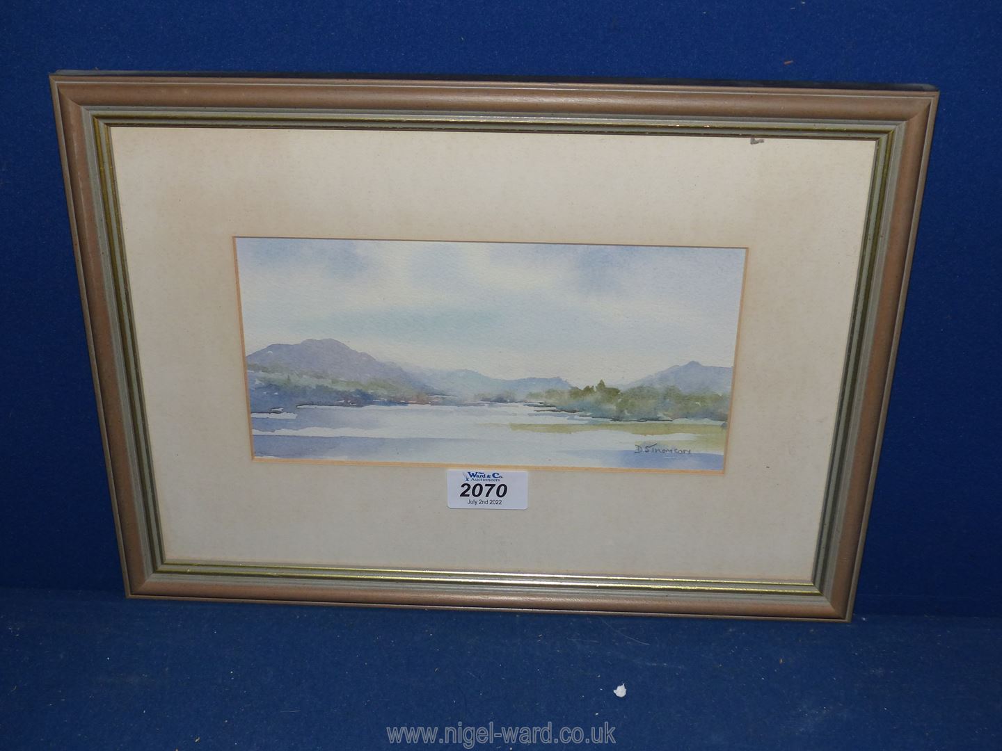 A framed and mounted watercolour depicting a river landscape with trees and hills in the distance,