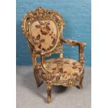 A large gold painted throne chair with floral upholstered seat, arm pads and studded surround on