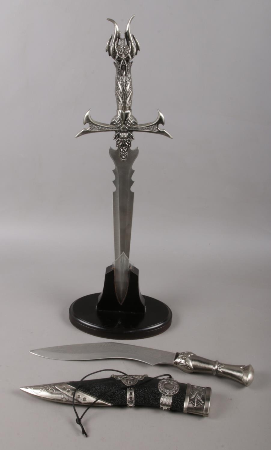 A large stainless steel dagger with mythical handle in stand along with a similar kukri knife in