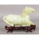 A Chinese carved jade figure of a recumbent horse, on hardwood stand. Length 14cm. Tip of ear