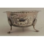 An Irish silver bowl, heavily embossed with flowers, buildings, figure and animal, raised on three