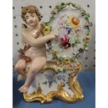 A 19th century porcelain ornament of a seated cherub by an oval floral encrusted plaque decorated