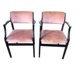 A pair of ebonised elbow chairs with padded backs and shaped arms above upholstered seats, raised on