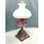 A Victorian cast iron oil lamp with cranberry glass reservoir and milk glass bowl shade framing
