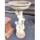 A composition stone birdbath with circular fluted bowl on column cast with three cherubs, raised