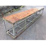 A long rectangular workbench on angle iron frame with six casters, the sheet metal top on plank