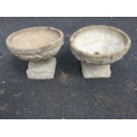 A pair of composition stone garden urns, the wide bowls moulded with scrolling on square plinths