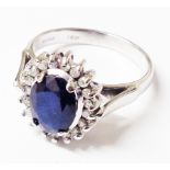 A marked 18K white metal ring, set with central oval sapphire within a diamond encrusted border -