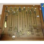 A wooden display board containing sixteen vintage brass toasting forks with various finial designs