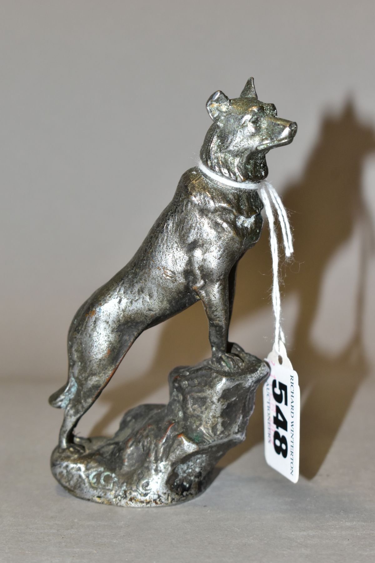 A CHROMED METAL CAR MASCOT IN THE FORM OF AN ALSATIAN STANDING WITH ITS FRONT LEGS ON A ROCK,