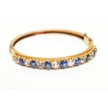 A YELLOW METAL GEMSET BANGLE, yellow metal hinged bangle set with seven oval cut blue sapphires
