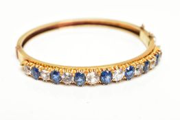 A YELLOW METAL GEMSET BANGLE, yellow metal hinged bangle set with seven oval cut blue sapphires
