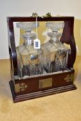 A MAHOGANY CASED TANTALUS WITH TWO DECANTERS bound with metal fittings, personalised plaque reads '