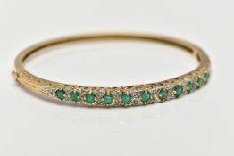 A 9CT GOLD, EMERALD AND DIAMOND HINGED BANGLE, designed with a row of eleven, claw set, circular cut