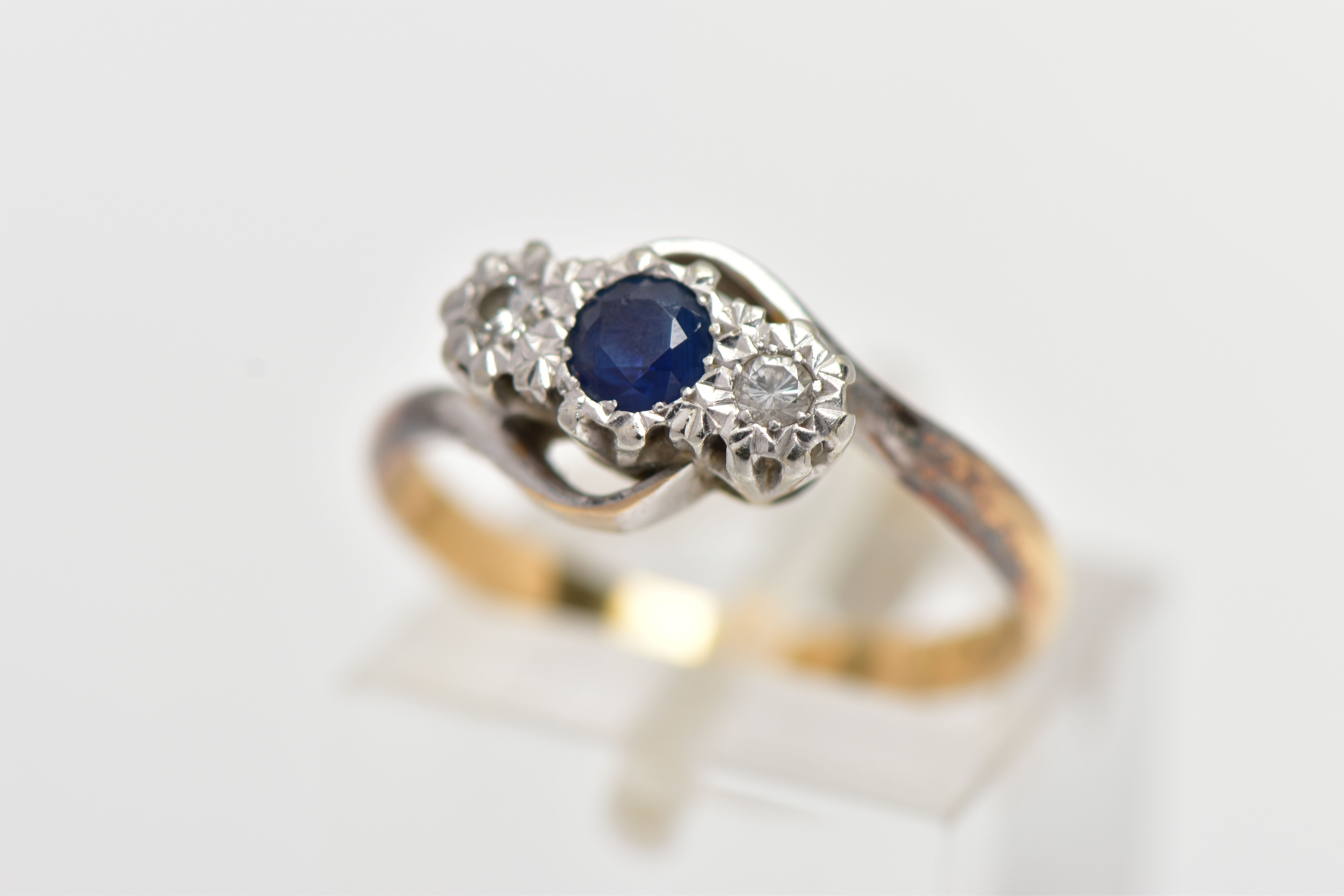 A YELLOW METAL CROSS OVER THREE STONE RING, designed with a central circular cut blue sapphire,