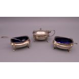 A silver three piece cruet set with blue glass liners and two spoons. Salts 3 cm high. 110.