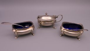 A silver three piece cruet set with blue glass liners and two spoons. Salts 3 cm high. 110.