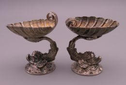 A small pair of Continental unmarked cast silver dolphin table salts, supporting shell bowls.