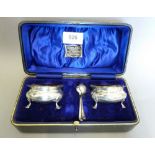 A pair of silver circular salts on shell and hoof feet with spoons, cased, Birmingham 1913