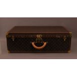A large Louis Vuitton suitcase, with a protective cover, 26cm H x 80cm W x 51.5cm D some water