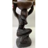 A heavy shaped dark wood sculpture of a nude lady kneeling whilst holding a bowl on her head.