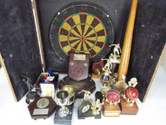 A dartboard in wall mountable cabinet, darts, baseball bat, various sporting trophies.