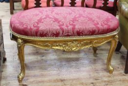 Large oval gilt framed stool in wine coloured classical floral fabric, 57cm by 114cm by 70cm.