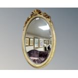 An oval-shaped bevel-edged mirror, in cream and gilt frame with ornate ribbon finial above,