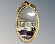 An oval-shaped bevel-edged mirror, in cream and gilt frame with ornate ribbon finial above,