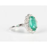 A white gold, treated emerald and diamond cluster ring, claw set with the oval cut treated emerald