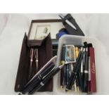 A tub containing a quantity of mostly ballpoint pens and a cased Parker fountain pen with ink