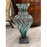 A large blue/green tinted fluted vase in wire work on an ornate square form metal base, 69cm tall