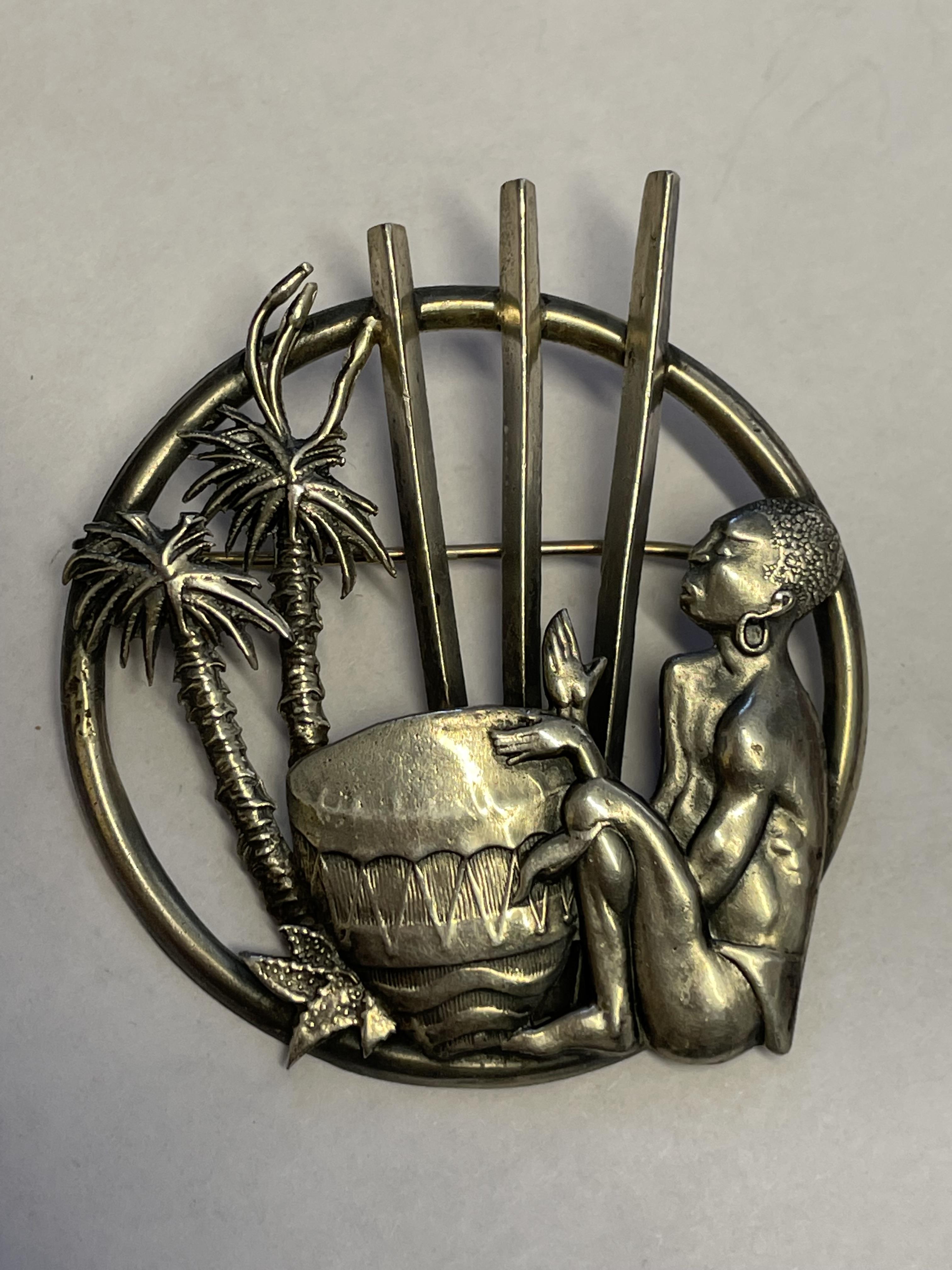 WHITE METAL BROOCH DEPICTING A MAN PLAYING A DRUM AMONGST PALM TREES