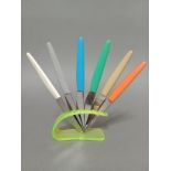 A vintage neon green knife rest with set of multi-coloured handled knives, the steel blades