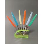 A vintage neon green knife rest with set of multi-coloured handled knives, the steel blades