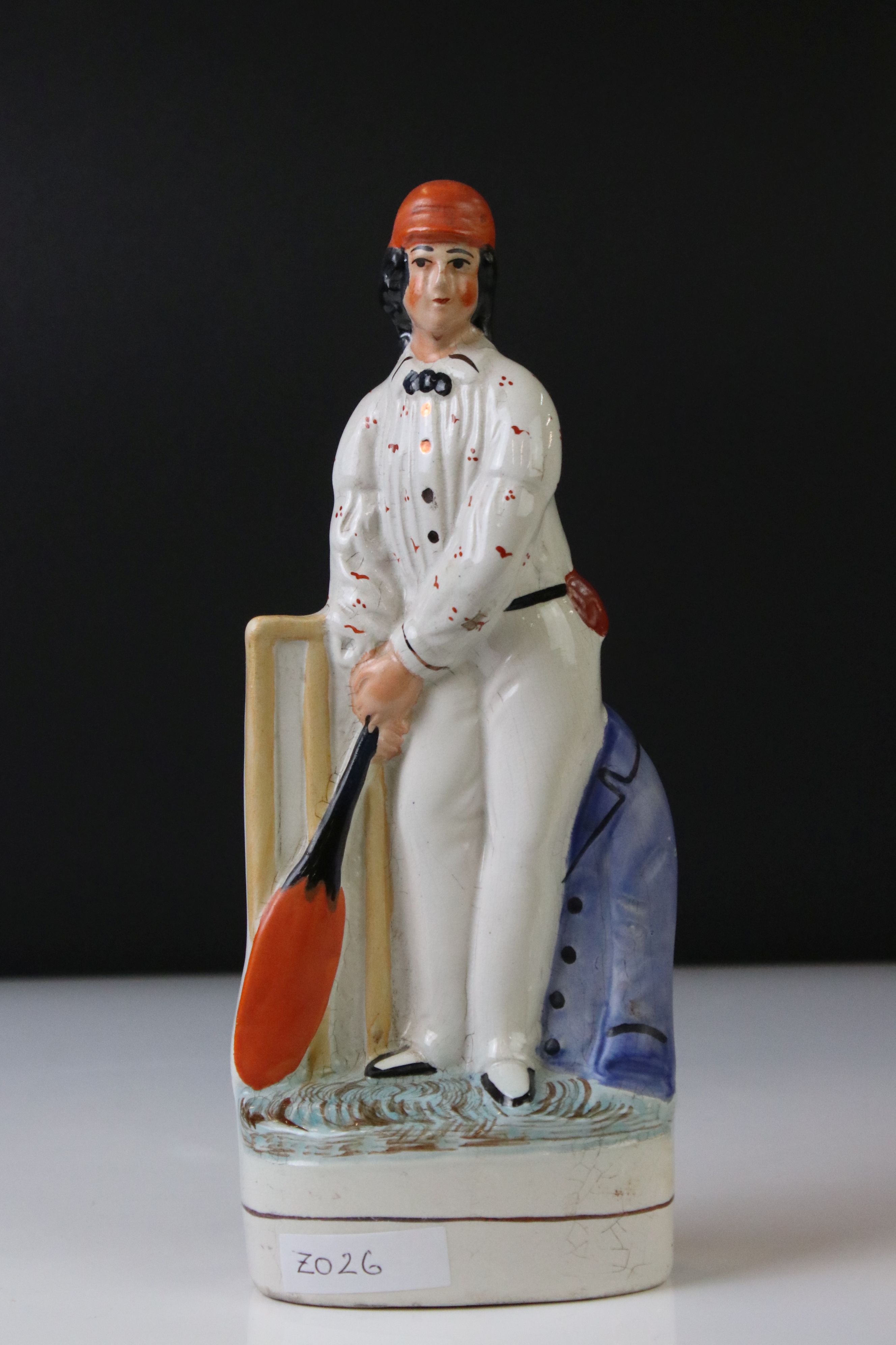 19th Century Staffordshire figure of a batsman wearing an orange cap, a cream spotted shirt, holding