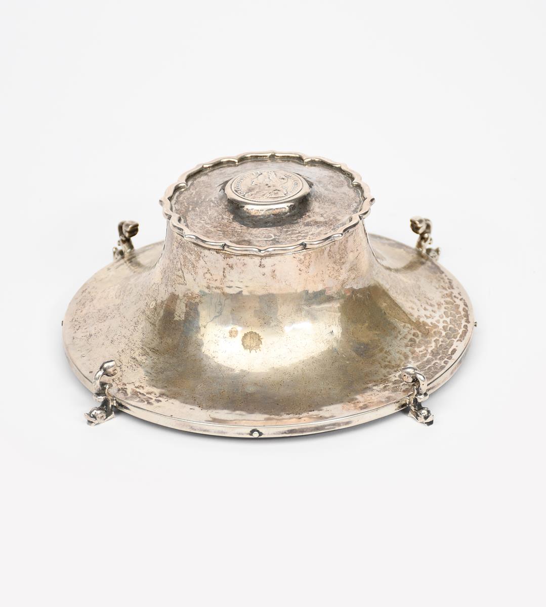 An A E Jones silver inkwell, circular form on dolphin feet, tapering hammered body, the hinged cover