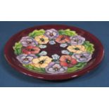 A Moorcroft plate with pansy garland decoration on a burgundy coloured ground, with impressed and