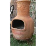 A weathered clay chimenea with sunburst mask detail, and raised on a simple iron work stand, 98 cm