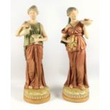 A pair of Royal Dux figures of serving girls dressed in neoclassical attire, one holding a ewer