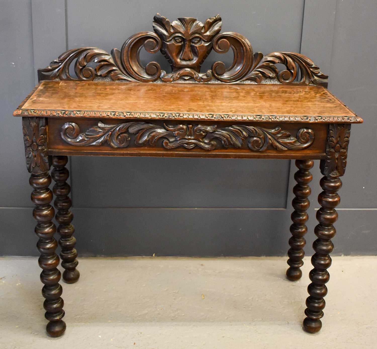 An oak 1930s silver table, the carved back depicting a face mask and scrollwork, with a frieze