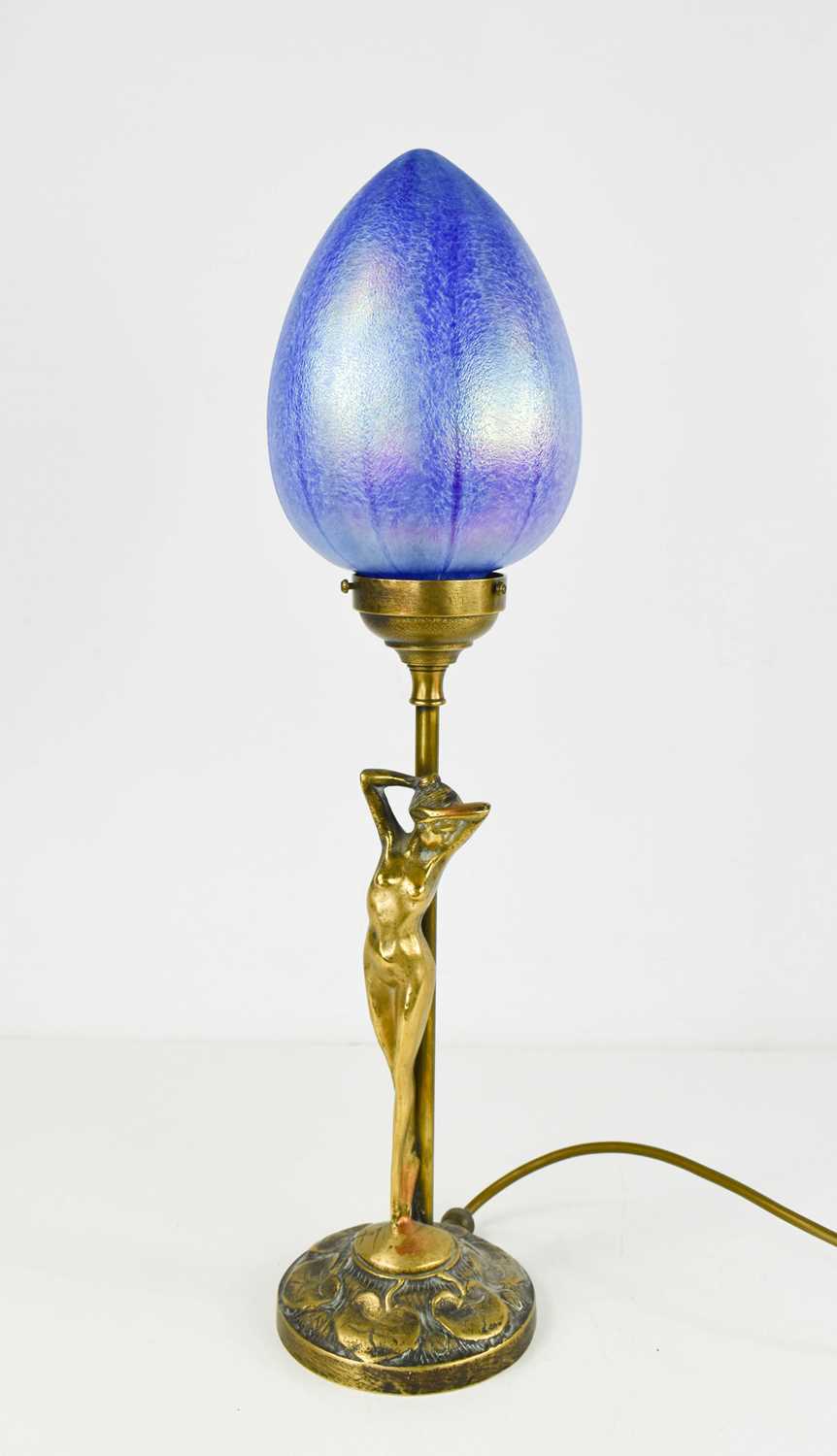 An Art Nouveau style brass table lamp in the form of a nude female, and having an iridescent glass