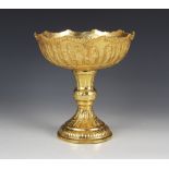 A gold coloured pedestal dish, the circular bowl embossed with a continuous frieze of figures in