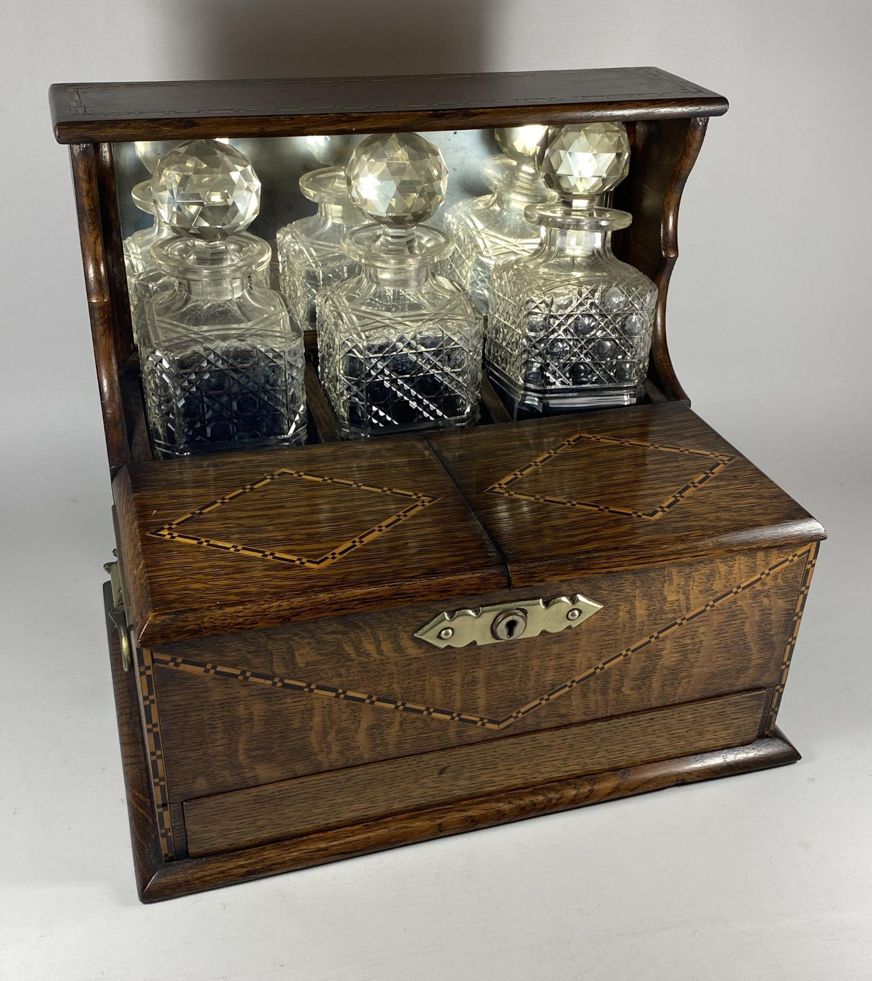 AN EDWARDIAN INLAID OAK THREE BOTTLE TANTALUS HOLDER WITH TWIN LIDDED COMPARTMENT AND LOWER DRAWER
