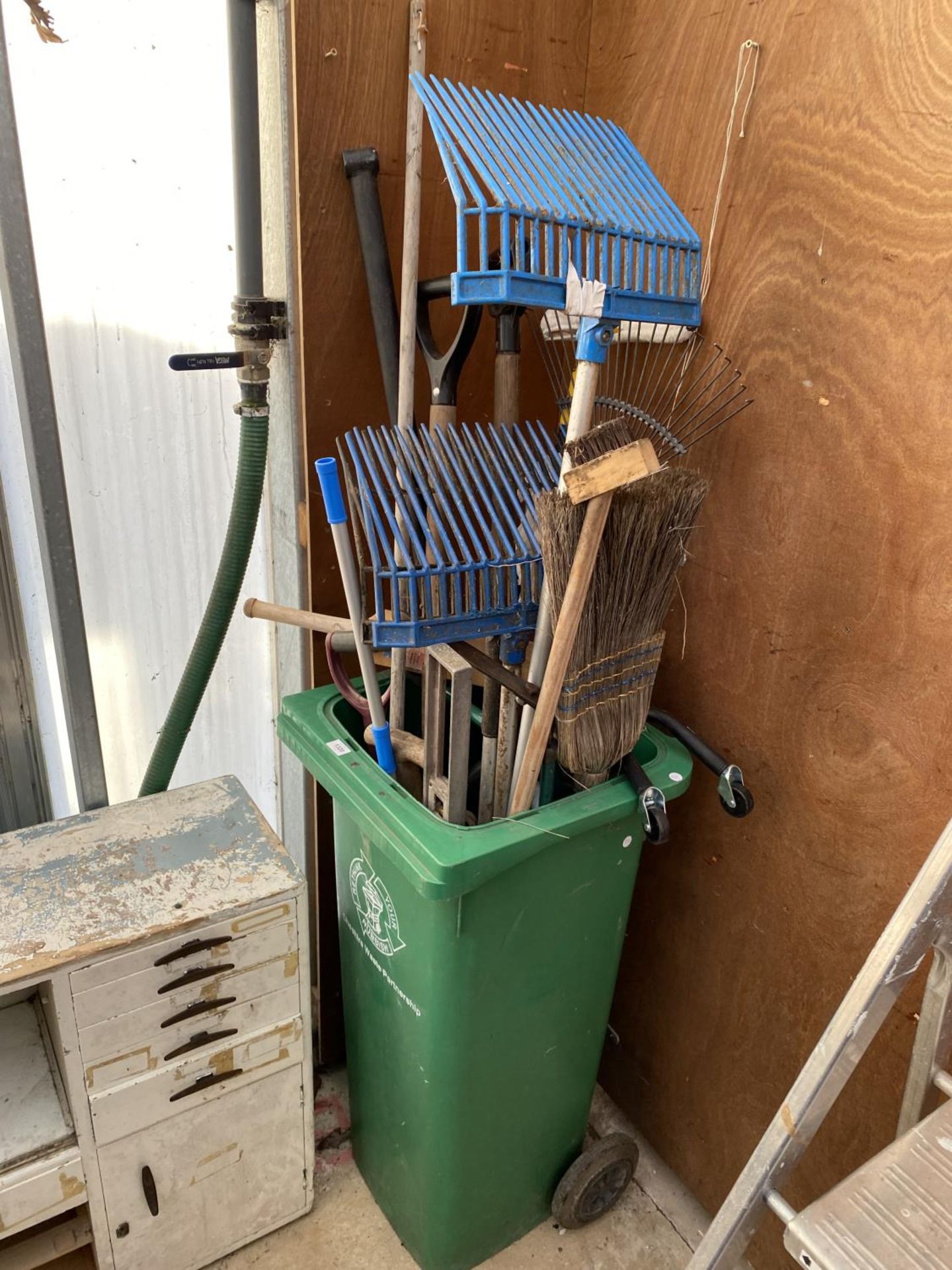 A WHEELIE BIN CONTAINING AN ASSORTMENT OF GARDEN TOOLS TO INCLUDE FORKS AND RAKES ETC