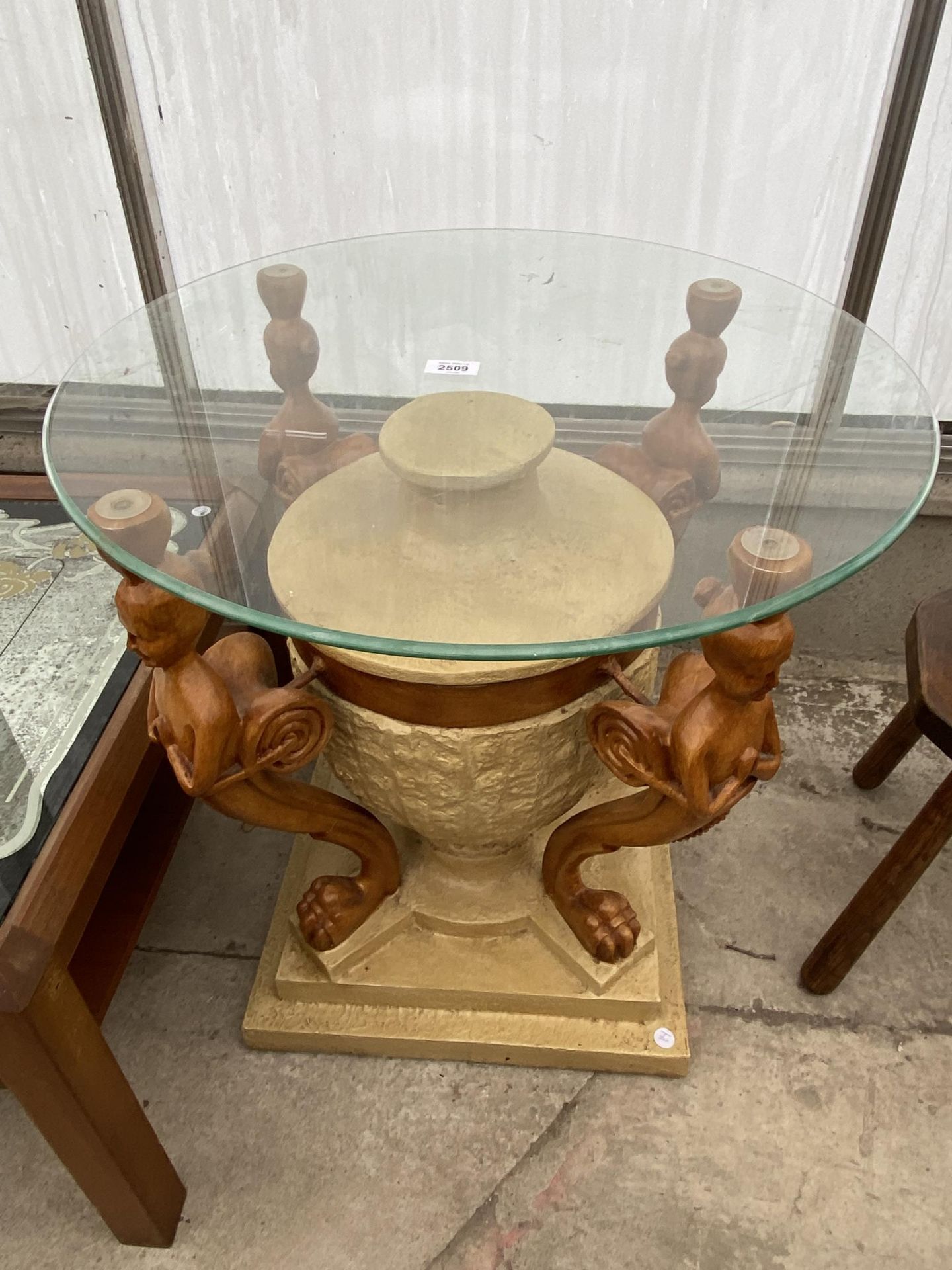 A RESIN LAMP TABLE ON URN SHAPED BASE SUPPORTED BY MYTHICAL FIGURES WITH GLASS TOP, 23.5" DIAMETER