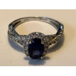 A 9 CARAT WHITE GOLD RING WITH A CENTRE BLUE GEMSTONE SURROUNDED BY CUBIC ZIRCONIAS WHICH ALSO TWIST