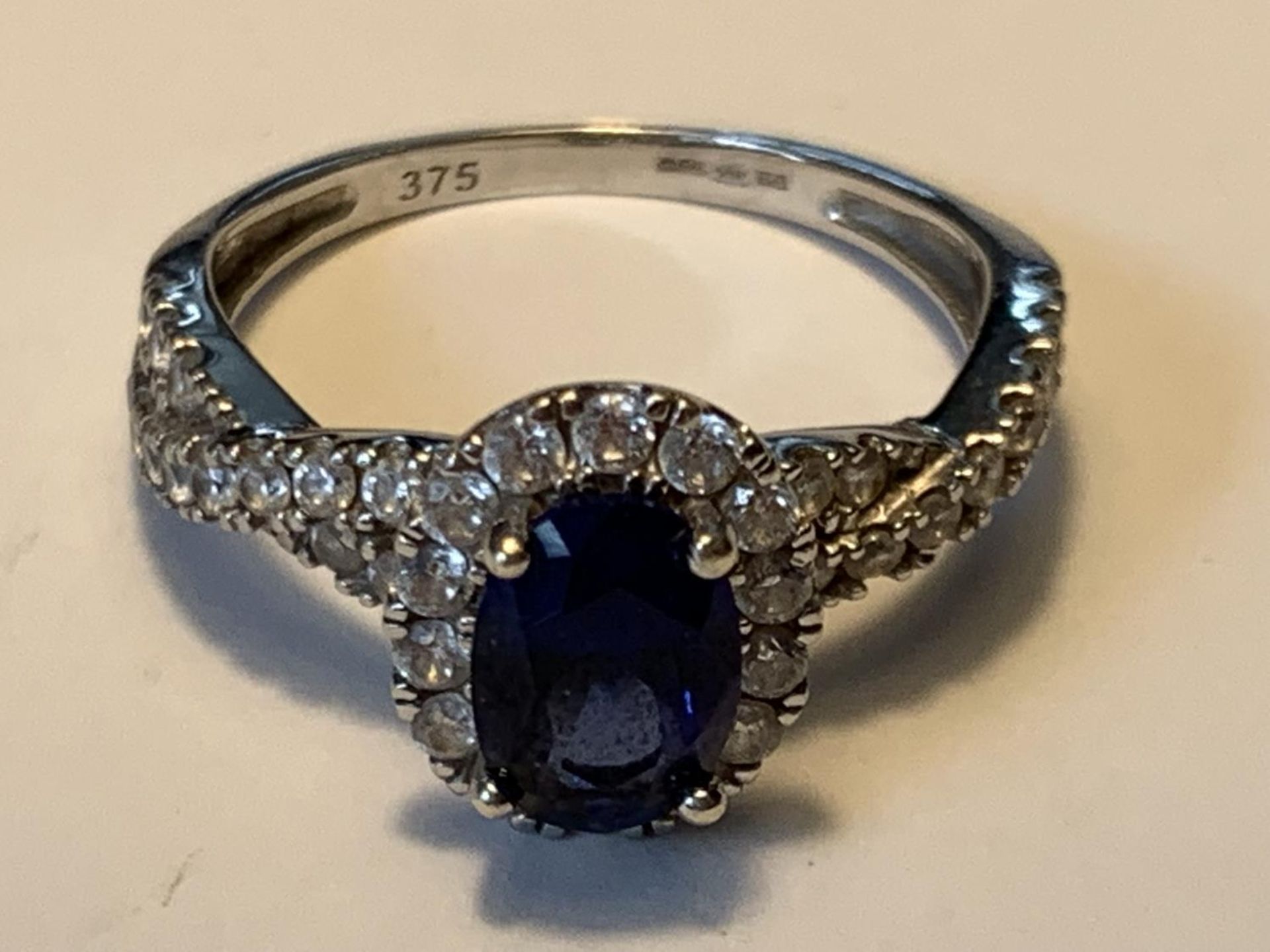 A 9 CARAT WHITE GOLD RING WITH A CENTRE BLUE GEMSTONE SURROUNDED BY CUBIC ZIRCONIAS WHICH ALSO TWIST