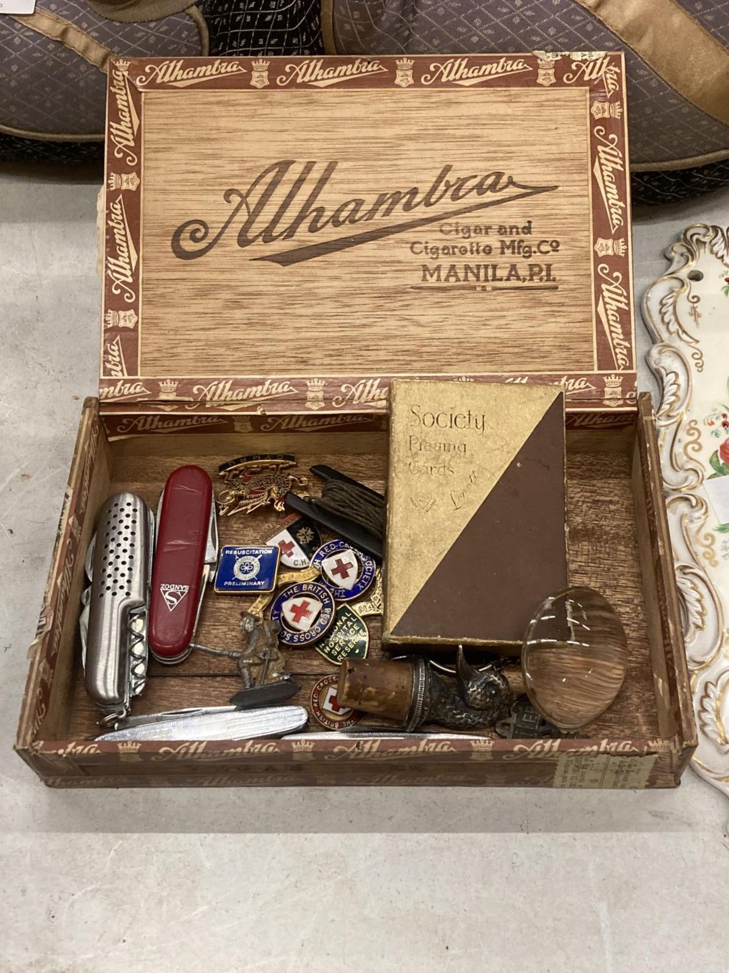 A CIGAR BOX CONTAINING VINTAGE PENKNIVES, RED CROSS BADGES, A BOTTLE STOPPER WITH WHITE METAL DUCK