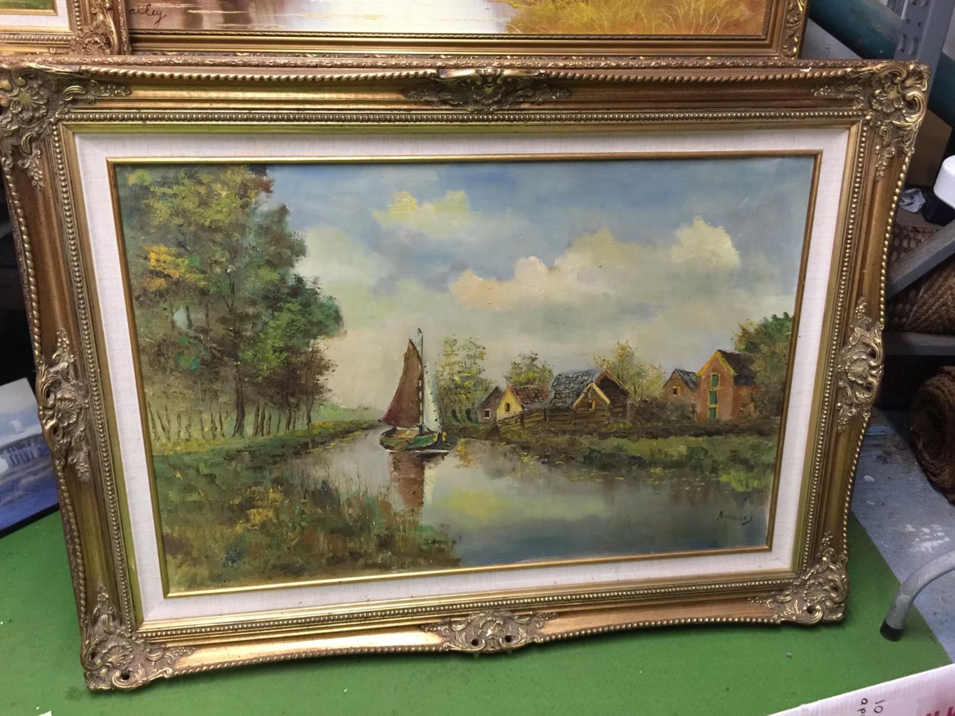 A LARGE GILT FRAMED OIL ON CANVAS OF A BOAT ON A STREAM FLANKED BY COTTAGES AND TREES, INDISTINCT